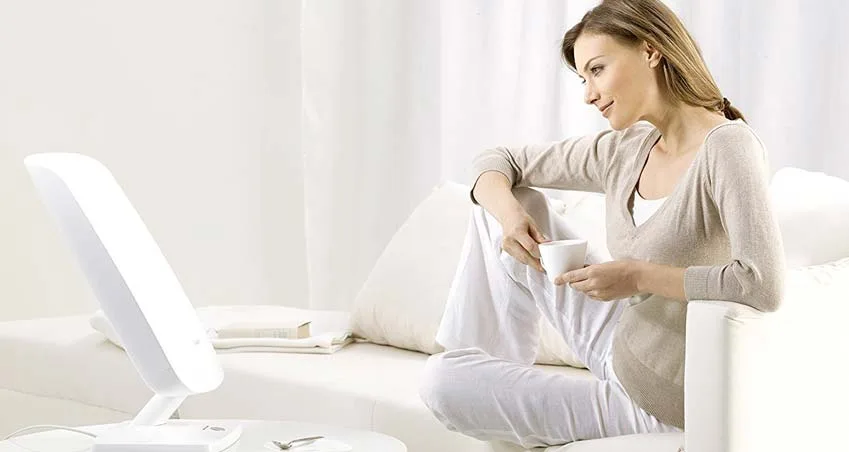 Eine Frau auf einer weußen VCouch. Vor ihr steht eine Tageslichtlampe von Beurer auf dem Couchtisch. Sie sieht sehr entspannt aus.