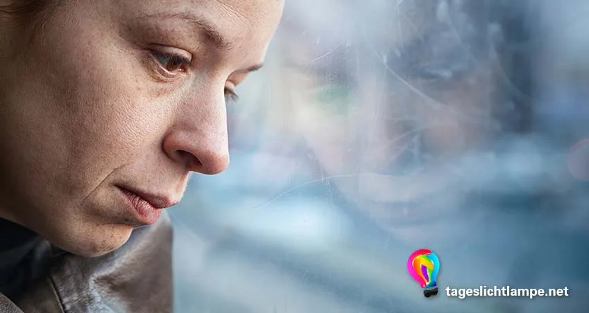 EIne depressive Frau sitzt am Fenster und guckt traurig - bei Depressionen hilft oft eine Tageslichtlampen Lichttherapie